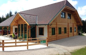 Eine Holzhütte mit grünem Dach und Fenstern, umgeben von Bäumen. Zwei Personen sitzen draußen an einem Tisch.