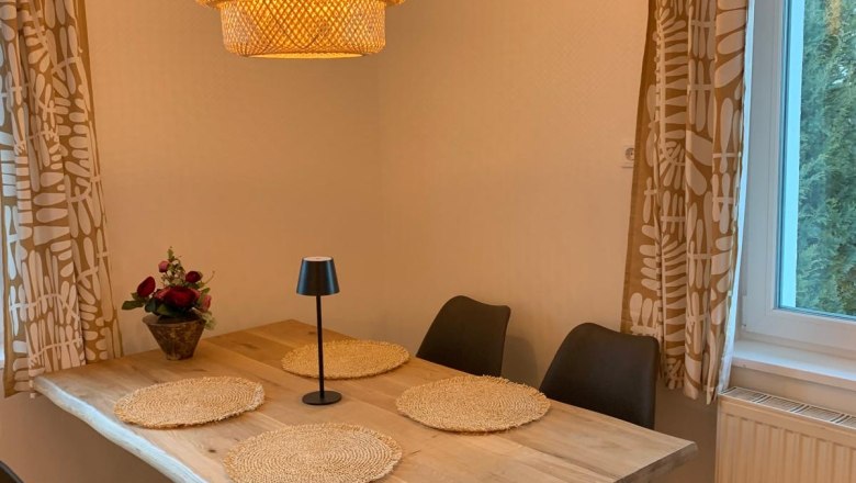 Essbereich mit Holztisch, vier schwarzen Stühlen, runder Lampe und Fenster mit Vorhängen.