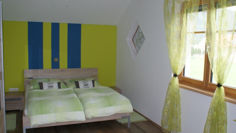 Schlafzimmer mit Doppelbett, grün-gelb gestreifte Wand, grüne Vorhänge, Holzboden.