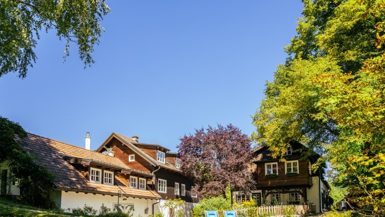 Ein traditionelles Landhaus mit Holzdach und blauen Liegestühlen im Garten, umgeben von Bäumen und blauem Himmel.