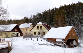 Verschneiter Bauernhof mit gelben Gebäuden und Holzscheune, umgeben von Wald.