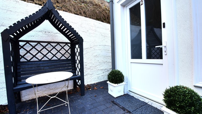 Eingangsbereich mit wei&szlig;er T&uuml;r, schwarzer Bank mit &Uuml;berdachung und kleinem Tisch.