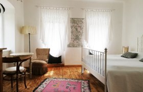 Helles Zimmer mit Holzboden, weißem Bett, zwei Fenstern mit Vorhängen, einem Tisch mit Stuhl und einem rosa Teppich.