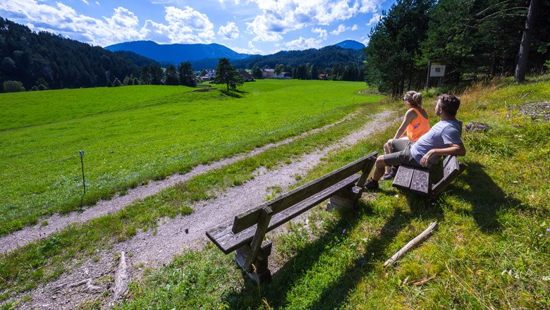 Zwei Personen sitzen auf einer Bank und blicken auf eine grüne Wiese mit Bergen im Hintergrund.