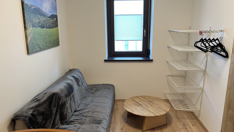 Kleines Zimmer mit Sofa, rundem Holztisch, Wandregal und Fenster.