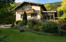 Ein zweist&ouml;ckiges Ferienhaus mit Balkon und Garten, umgeben von B&auml;umen und Wiese.