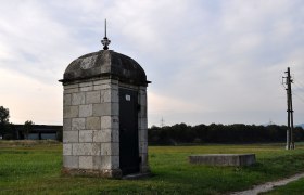 Steinbauwerk auf einer Wiese mit Strommast im Hintergrund.