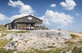 Fischerhütte auf dem Schneeberg mit Wanderern und blauem Himmel.