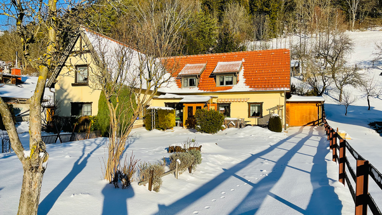 Schneebedecktes Haus mit rotem Dach und Garten, umgeben von Bäumen und Zaun, bei sonnigem Wetter.