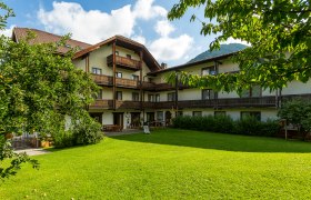 Ein traditionelles Hotel mit Holzbalkonen und einem gepflegten Garten im Vordergrund, umgeben von Bäumen und Bergen im Hintergrund.