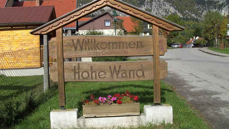 Holzschild mit der Aufschrift 'Willkommen in der Gemeinde Hohe Wand' vor einer Berglandschaft.