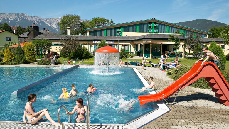 Freibad mit Rutsche und Pilzbrunnen, umgeben von Bergen.