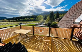 Holzbalkon mit Tisch, Blick auf Wiesen und Berge, Sonnenstrahlenmuster auf dem Boden.