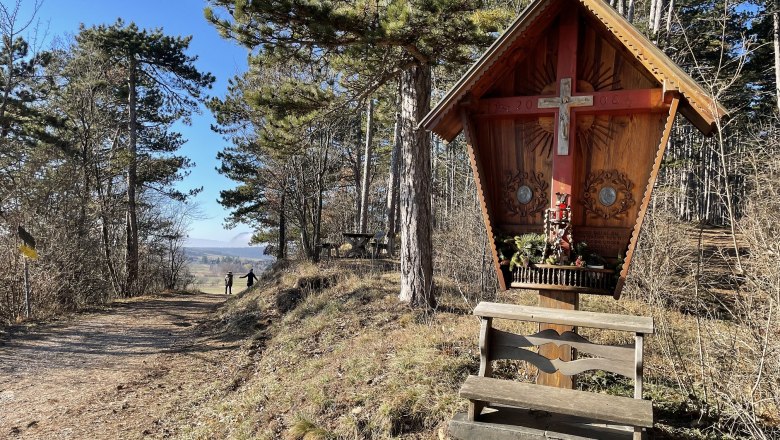 Holzkreuz mit Bank in einem Waldgebiet, sonniger Tag.